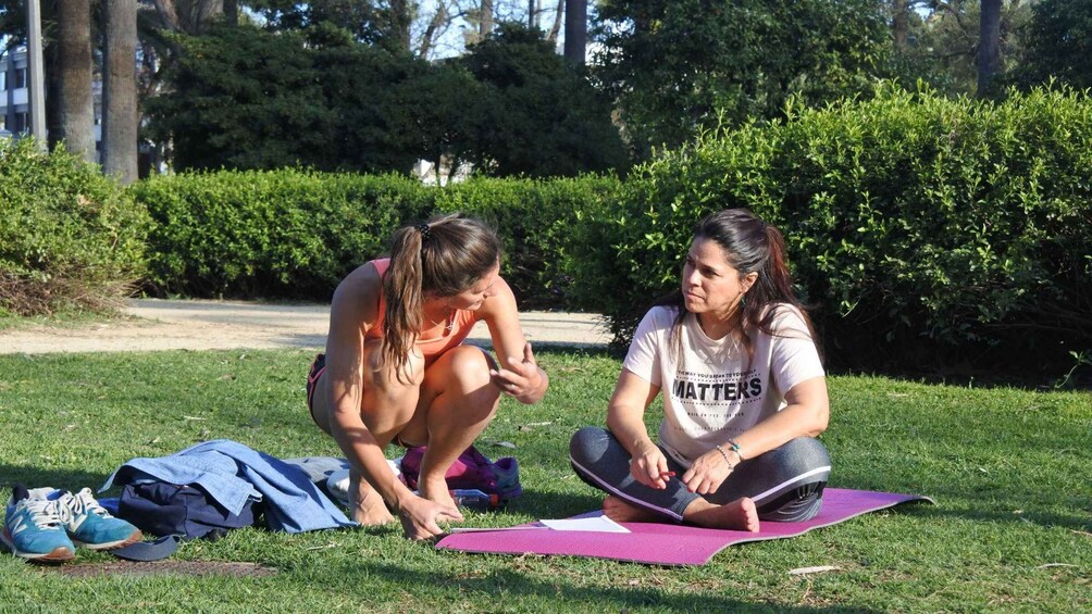 Two people sitting on a purple yoga mat outdoors, one on a blue mat and the other on a pink mat, with a blue bag and shoes on the ground. 3