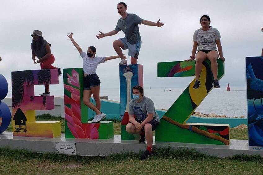People standing on large, colorful letters outdoors with a body of water in the background. 2