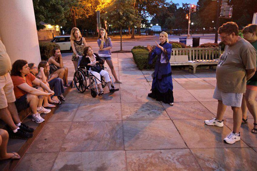 A group of people sitting and standing on a sidewalk, some in wheelchairs, engaged in a discussion with a person in a blue robe. 5
