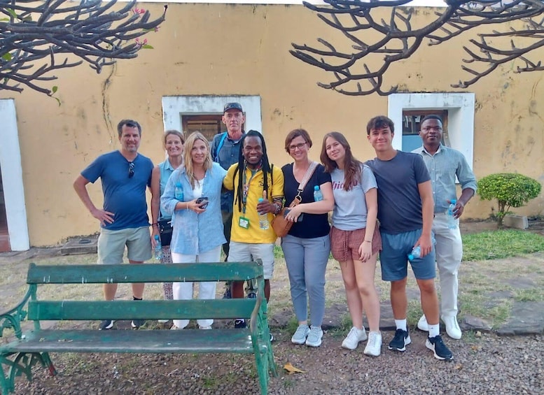 A group of people standing in front of a building with two windows and a bench in the foreground. 1