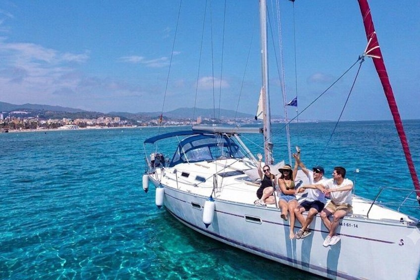 A sailboat with people on deck, clear blue water, and a distant shoreline. 1