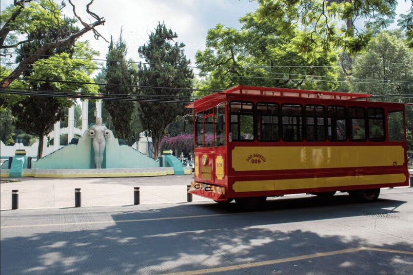 A red and yellow trolley on a street with trees and a fountain in the background. 1