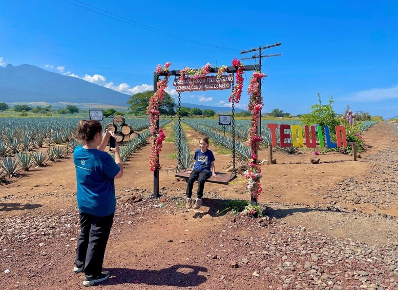 A rural landscape with a person taking a photo, a swing set, and a sign with 'TEQUILA' written on it. 4