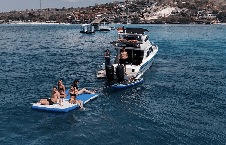 A boat with people on inflatable mats in the water, a docked boat, and houses in the background. 1