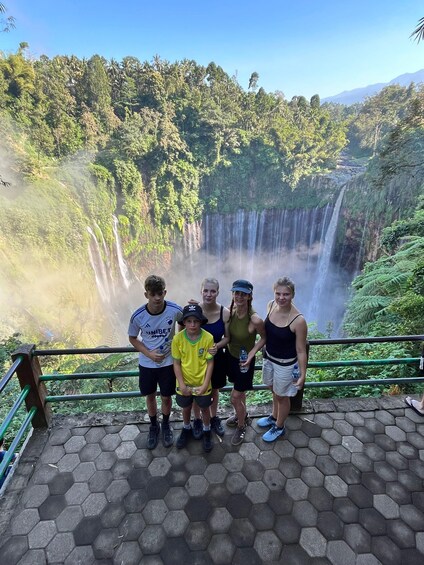 A group of people standing on a paved area with a hexagonal pattern, overlooking a waterfall surrounded by lush greenery and trees. 2