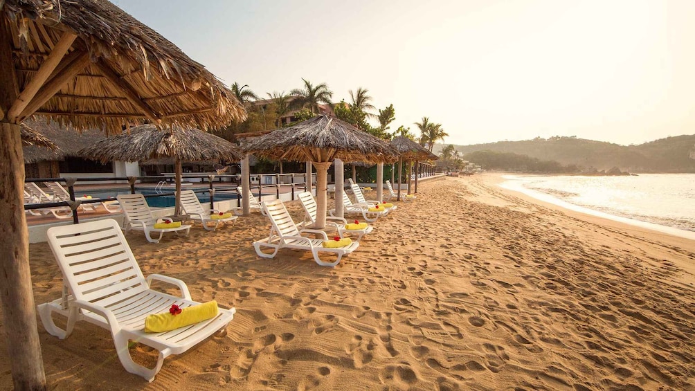 A beach with white lounge chairs and thatched umbrellas. 1