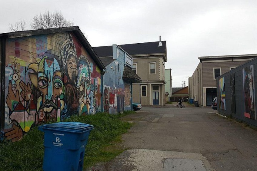A street with graffiti on a building, a blue recycling bin, and a cyclist in the distance. 3