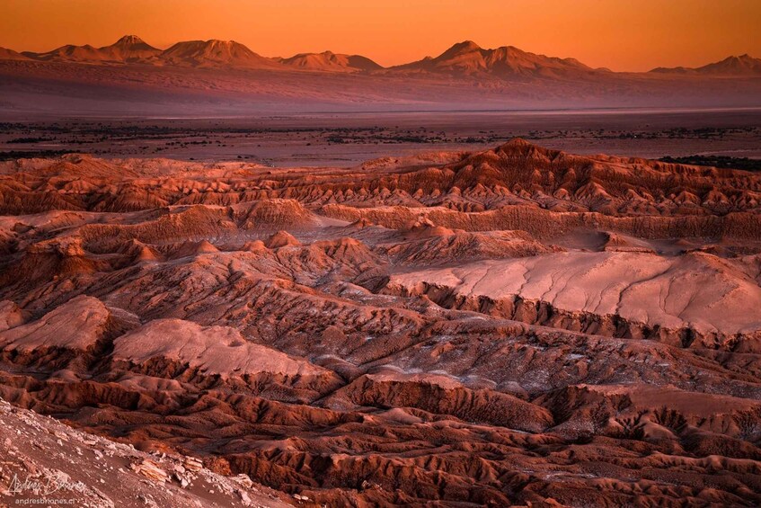 Un paysage désertique accidenté, avec des collines érodées et des montagnes lointaines, sous un ciel embrasé par le coucher du soleil. 2