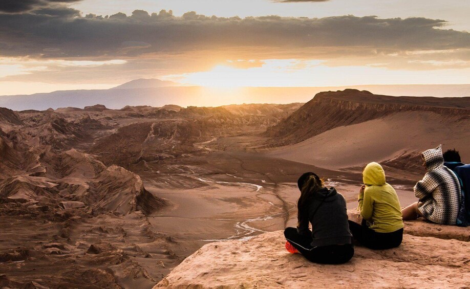 Trois personnes sont assises sur un rocher, d’où elles dominent un paysage désertique avec une rivière et des montagnes à l’horizon. 4