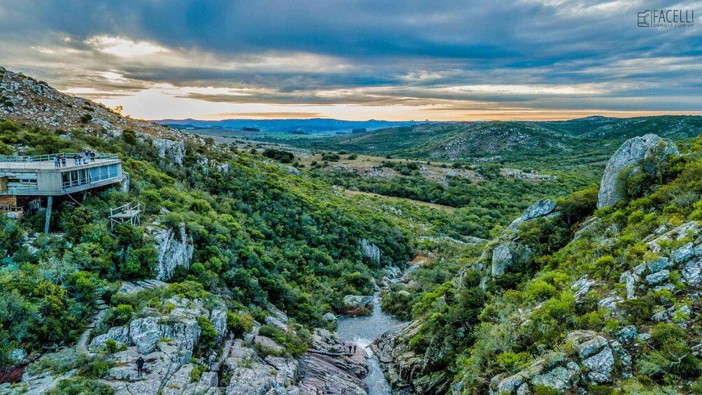 A rocky gorge with a river, a wooden viewing platform, and a distant mountain range under a cloudy sky. 2