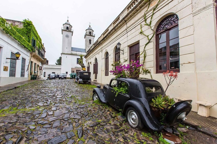 A cobblestone street with a vintage car parked on the side, a church with two towers, and buildings with ivy and flowers. 4