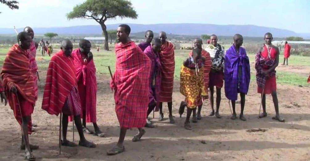 A group of people in traditional attire walking in a line on a dirt path with a tree and mountains in the background. 1