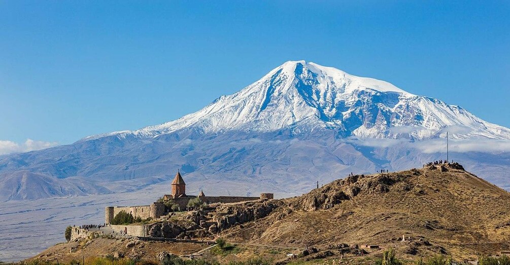 A snow-capped mountain, a historic church, and a rocky hill with people. 1