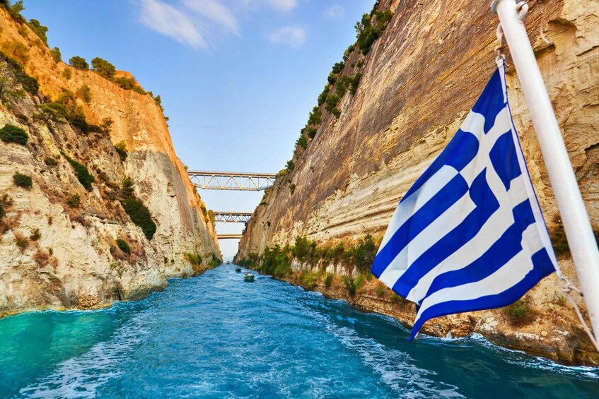 A narrow waterway flanked by steep cliffs, a bridge in the distance, and a Greek flag. 5
