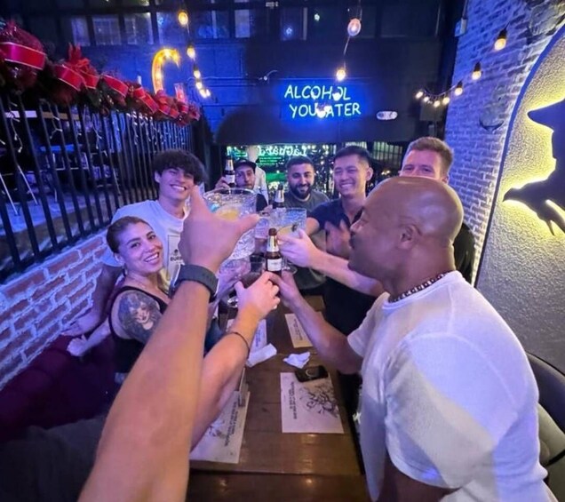 A group of people toasting with drinks at a bar with a neon sign reading 'ALCOHOL YOU WATER.' 1