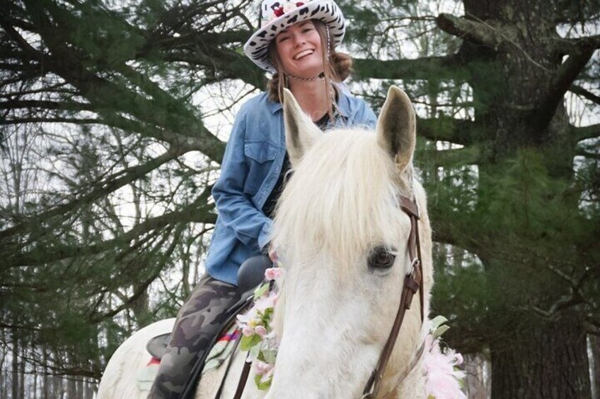 Een persoon rijdt op een witte paard met een zadel versierd met bloemen, draagt een helm en een jeansjas, in een bebost gebied. 5