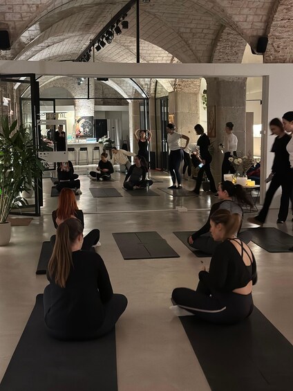 A group of people sitting on yoga mats in a spacious indoor area with a high ceiling and brick walls. 2