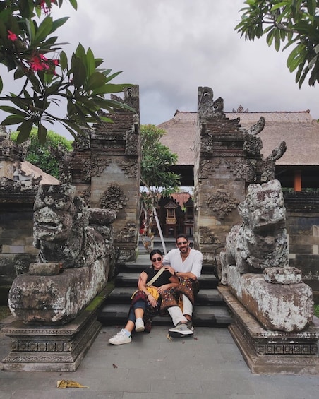 A couple sitting on stone steps in front of a temple with intricate carvings and a thatched roof. 3