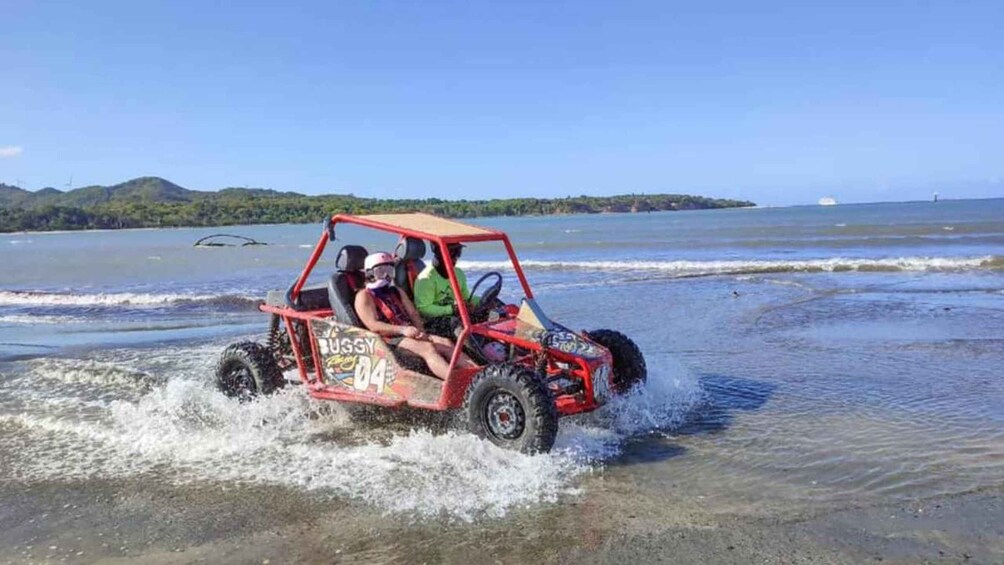 A red buggy with 'BUGGY 04' and 'RUGGY' on the side, driven by two people on a beach with waves and a distant island. 2