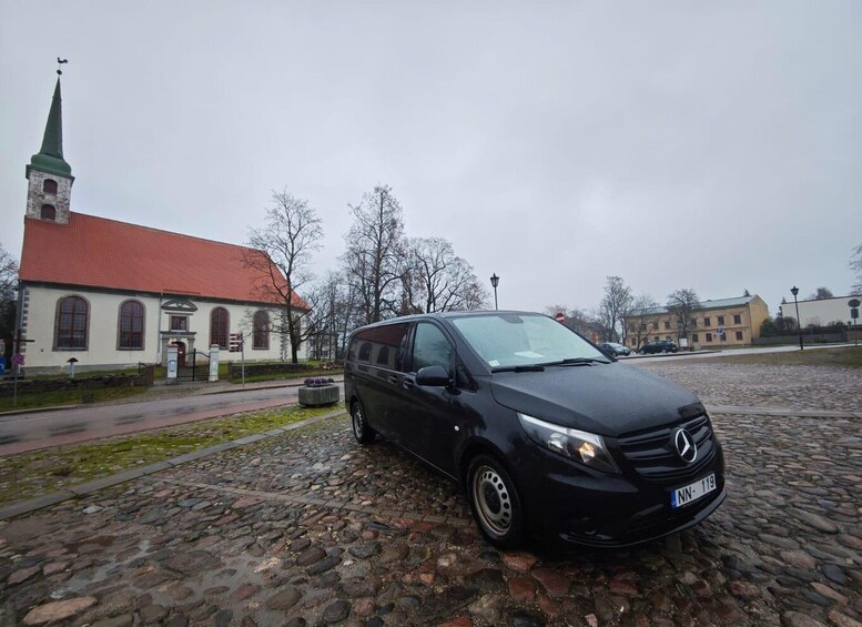 A black car parked on a cobblestone street in front of a church with a red roof. 2