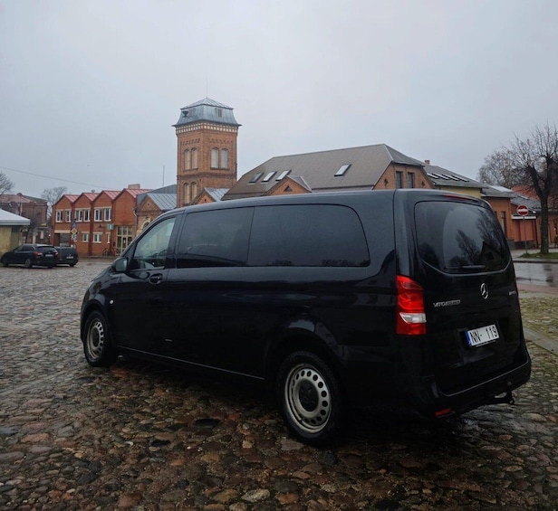 A black van parked on a cobblestone street with a brick building and a tower in the background. 1