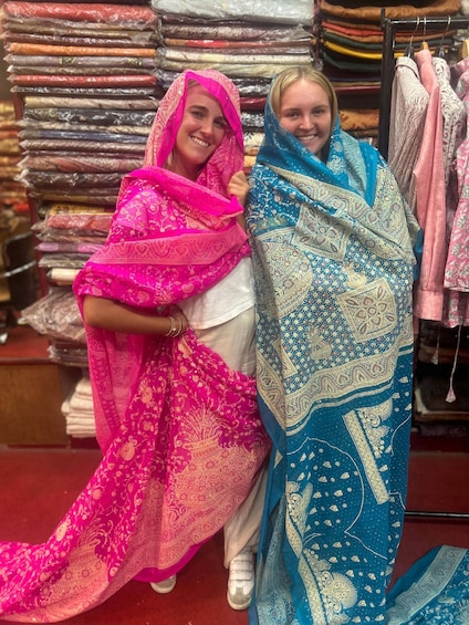 Two people wrapped in colorful fabrics, standing in front of a stack of folded fabrics. 2