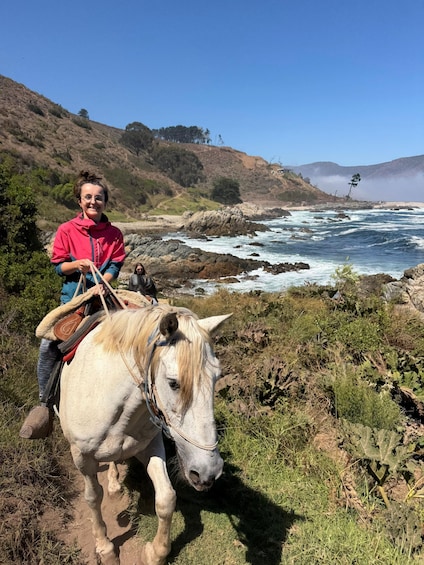 A person riding a white horse along a coastal trail with rocky ocean waves in the background. 1