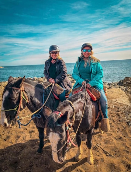 Two people riding horses on a rocky beach with the sea in the background. 2