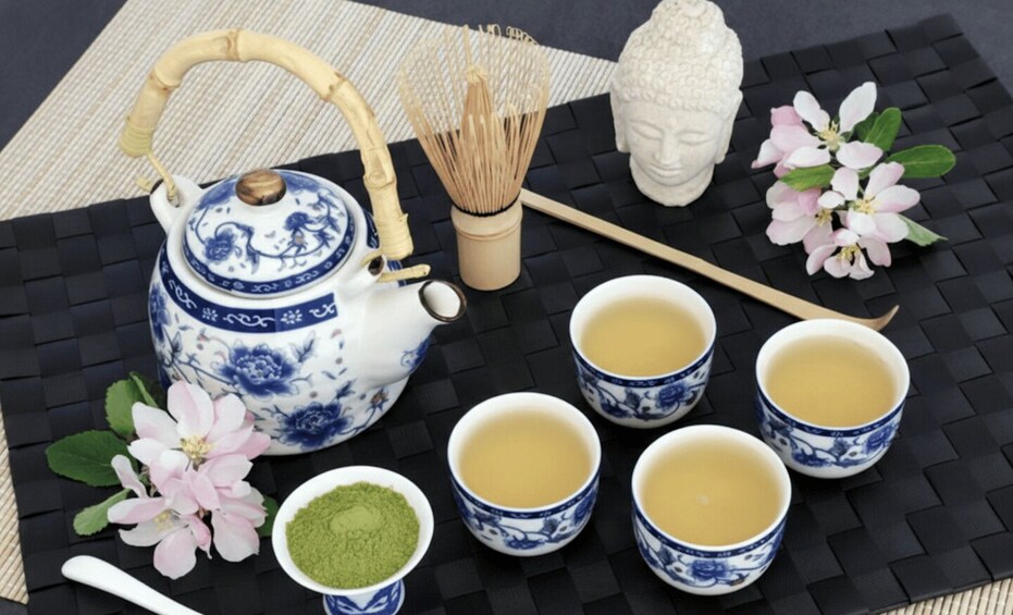 A traditional Japanese tea set with a teapot, cups, and a whisk, accompanied by flowers and a Buddha statue. 1