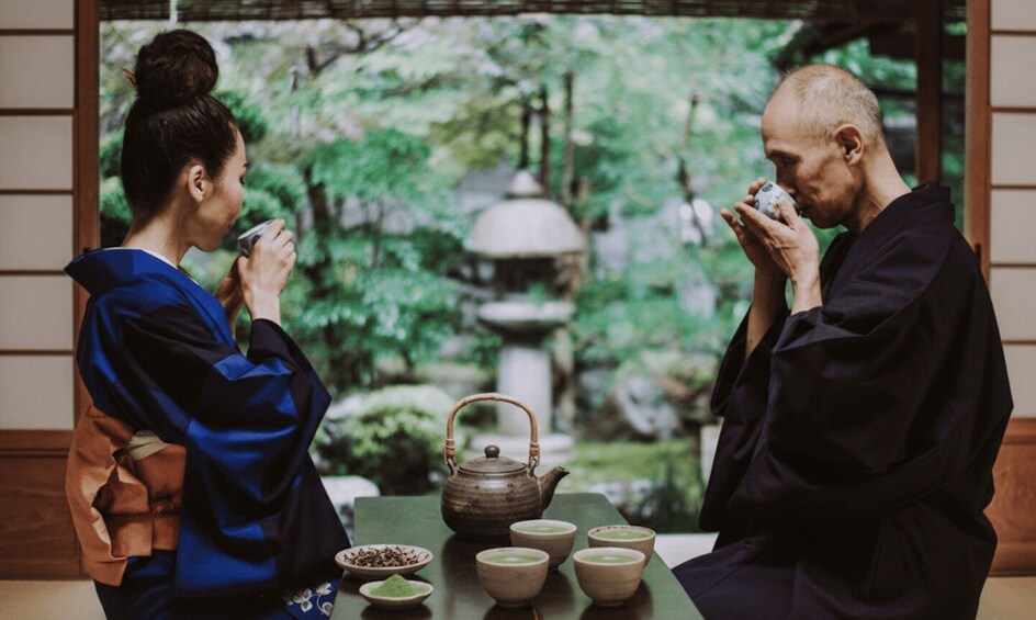 Two people in traditional attire sitting at a table with a teapot and cups, in a room with a view of a garden. 2