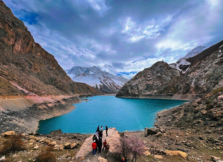 A group of people standing by a turquoise lake surrounded by mountains. 2