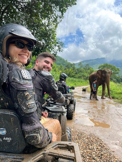 Two people in protective gear on an ATV, with an elephant and a dog in the background. 4
