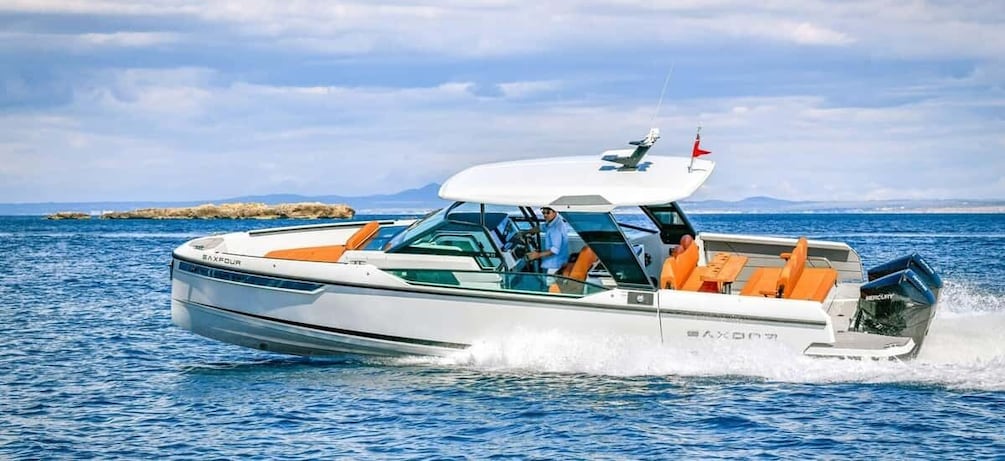 A white speedboat with orange seating and a red flag on top, moving on a calm sea with a rocky shoreline and mountains in the background. 1