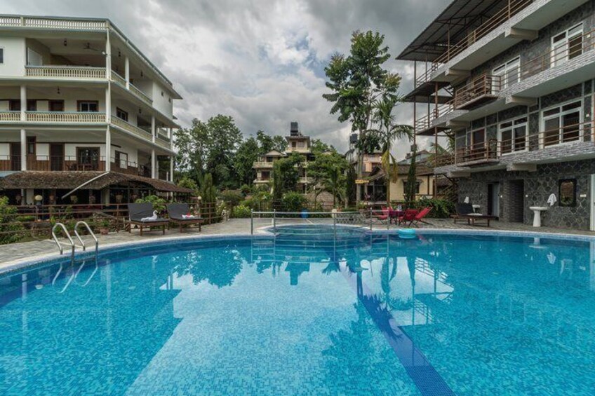 A swimming pool with lounge chairs, a restaurant, and multi-story buildings in the background. 3