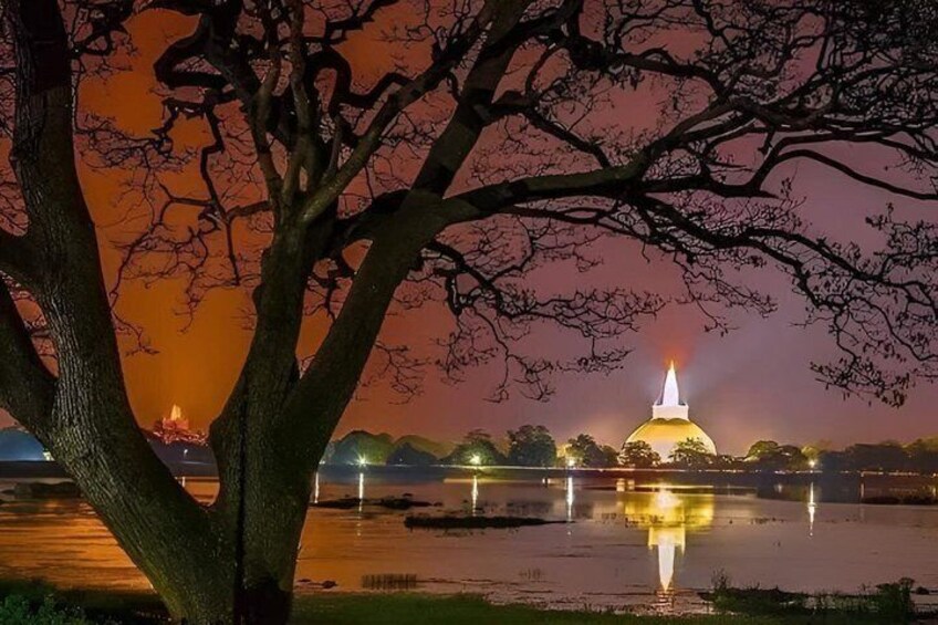 A night scene with a lit dome structure reflecting on water, surrounded by trees and distant lights. 1
