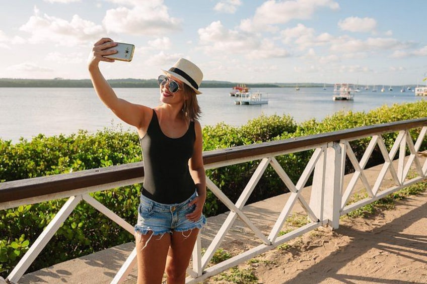 A person taking a selfie with a smartphone, wearing a black tank top, denim shorts, and a hat, standing on a concrete path with a railing, overlooking a body of water with boats. 1