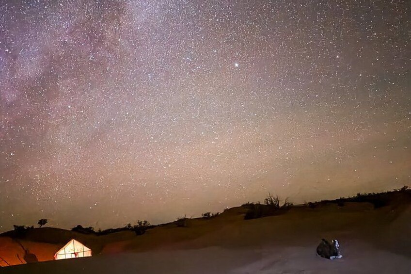 A starry night sky over a hilly landscape with a small illuminated structure and a person sitting on the ground. 3