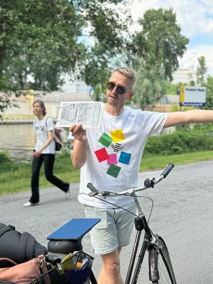 A person holding a map, wearing a white t-shirt with colorful squares, standing next to a bicycle on a paved path. 4