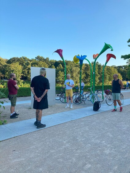 People standing outdoors near a white structure, with bicycles and green sculptures in the background. 2