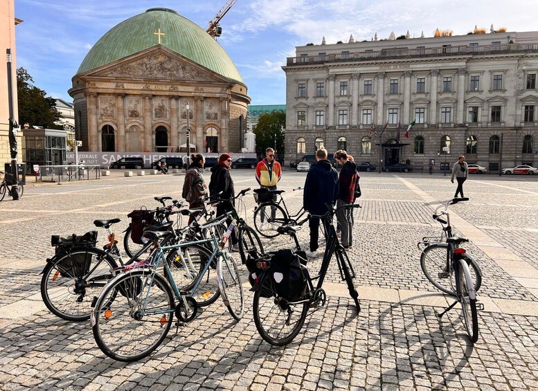 A group of people standing in a square with bicycles parked in front of a historic building with a green dome. 1