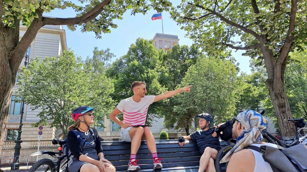 A group of people sitting on a bench in a park with trees and a building in the background. 5
