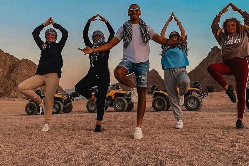 Cuatro personas bailando en un desierto con vehículos todo terreno al fondo. 3