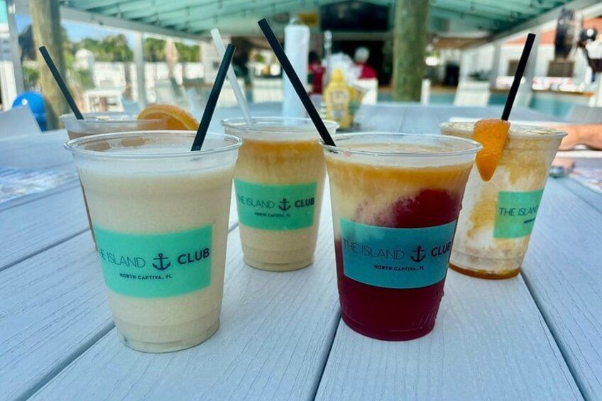 Tres bebidas heladas con pajitas y una rodaja de naranja a la derecha, identificadas como 'The Island Club', sobre una mesa de madera blanca. 5