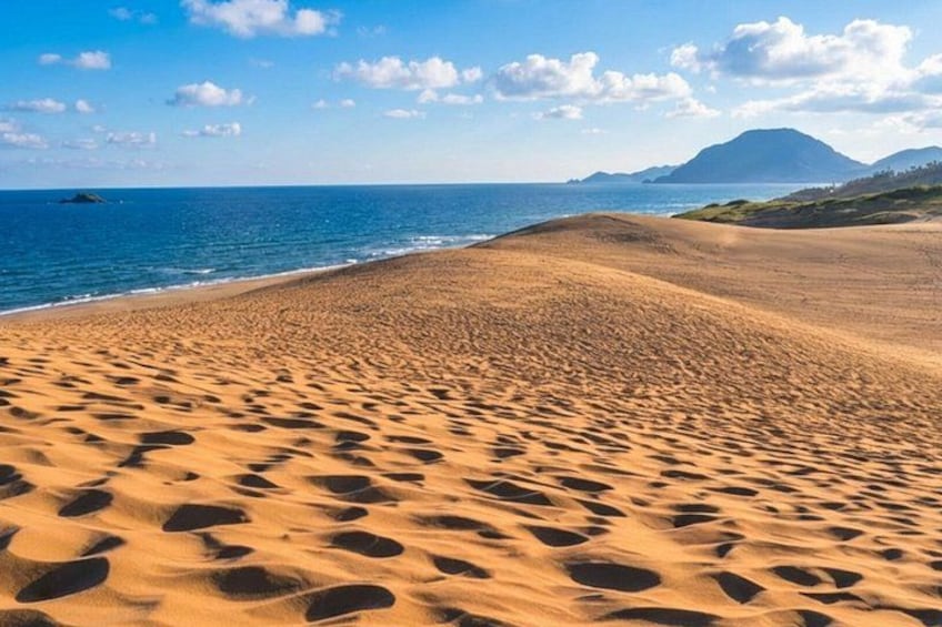 A sandy beach with footprints, a calm sea, and a mountain in the background. 1