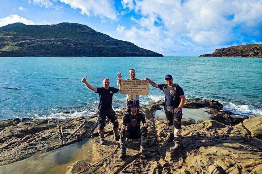 Three people on a rocky shore with a sign, a body of water, and a distant mountain. 4