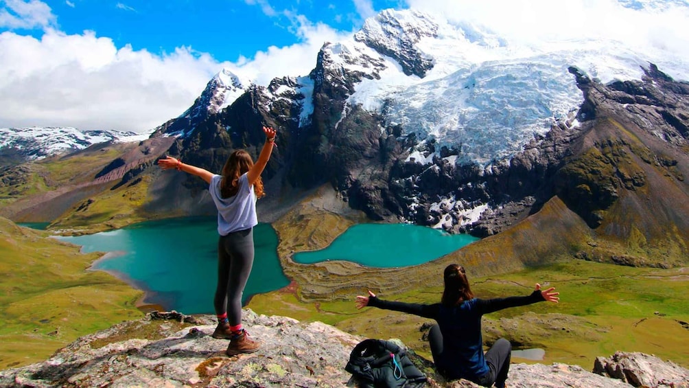 Two people standing on a mountain peak with arms outstretched, overlooking turquoise lakes and snow-capped mountains. 1