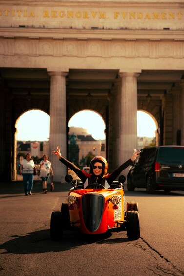 Une voiture ancienne, conduite par un chauffeur portant un casque, roule sous un pont orné de colonnes et d’arches. 2