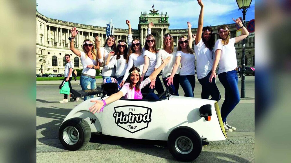 Un groupe de personnes posant devant un grand bâtiment historique, à côté d’une voiture blanche de la série Hotrod Tour. 5