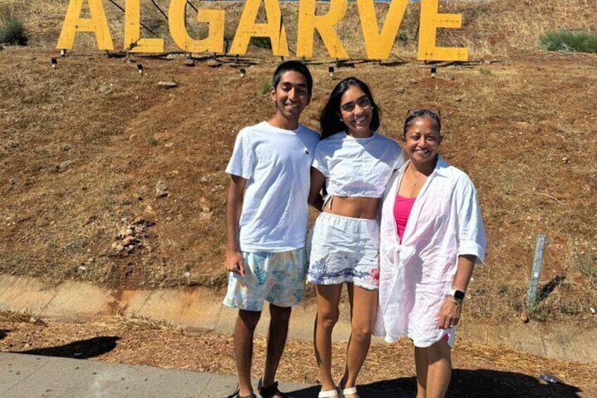 Tres personas de pie frente a un letrero amarillo grande que dice 'ALGARVE'. 3