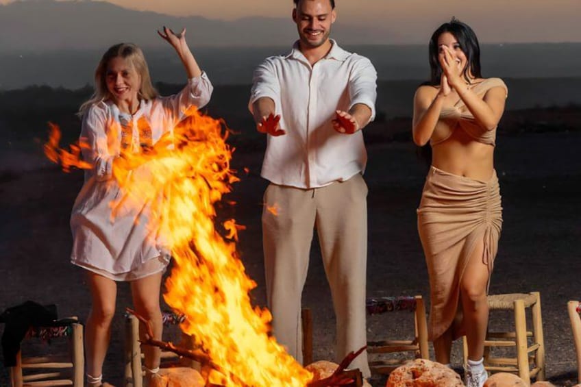 Three people standing near a fire with a mountainous landscape in the background. 5